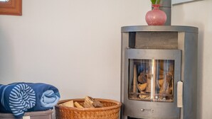 Una habitación de esquina con estufa de leña, un cesto de leña y una manta enrollada sobre un baúl.