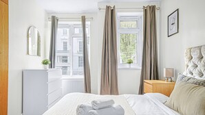 A hotel room with a bed, a dresser, a nightstand, a window with curtains, and a view of buildings outside. - Paddington Station 2BR City Apartment (London)