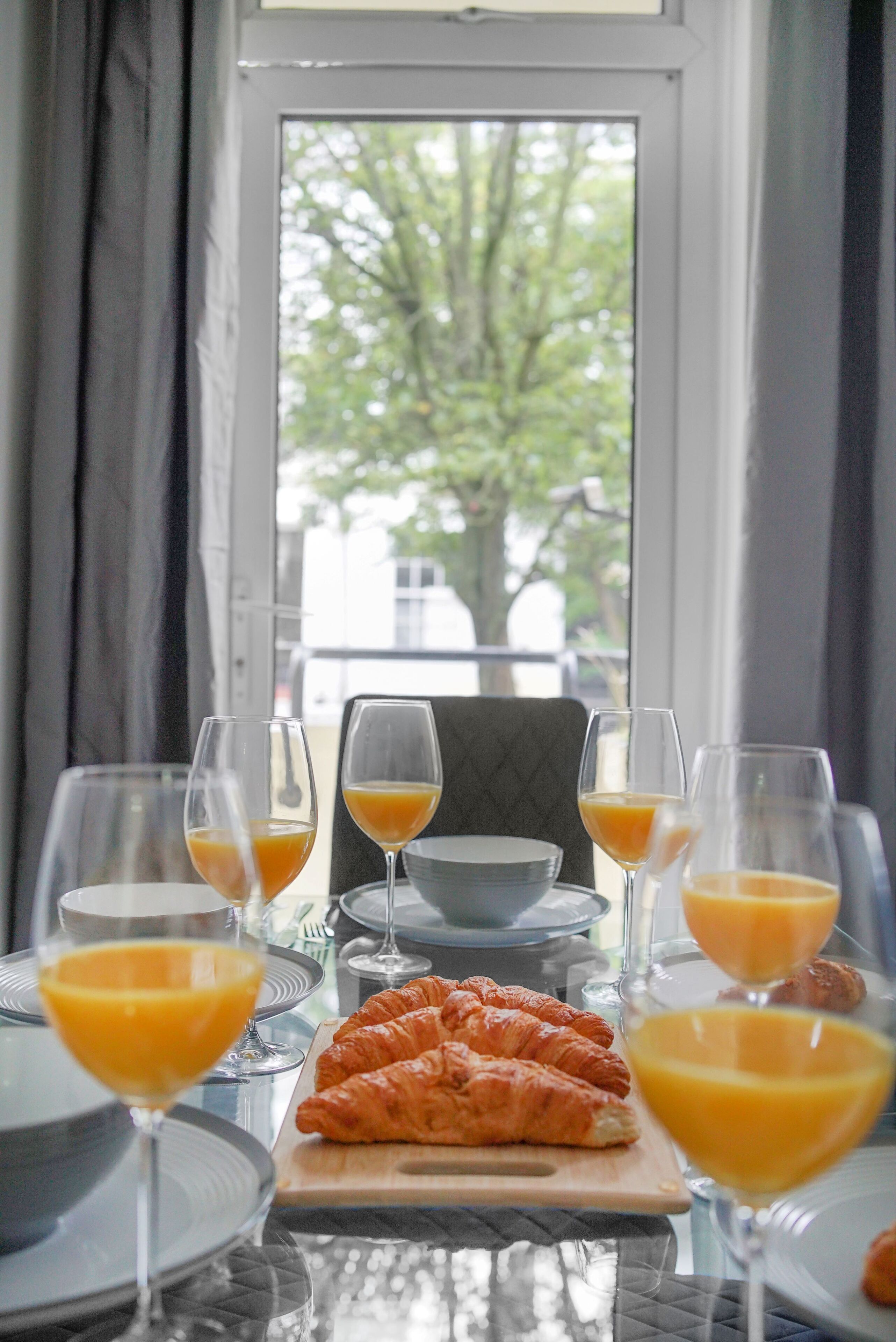 A breakfast table with croissants, orange juice, and glasses, set against a window with a view of trees.