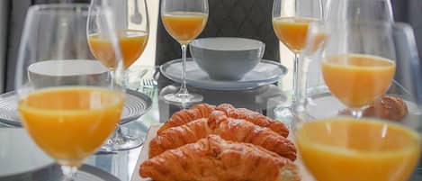 Una mesa de desayuno con cruasanes, zumo de naranja y vasos, frente a una ventana con vistas a árboles.