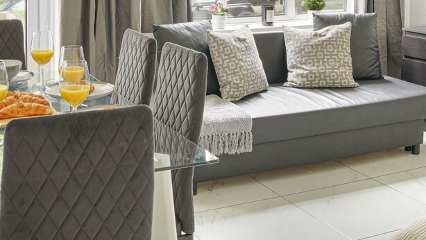 A modern living room with a grey sofa, quilted chairs, a dining table with orange juice, and large windows with curtains. - Paddington Station 2BR City Apartment (London)