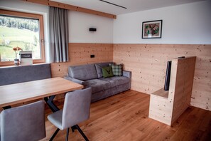 A modern room with a wooden floor, a grey sofa with cushions, a wooden table, and a window with a view of a green landscape.