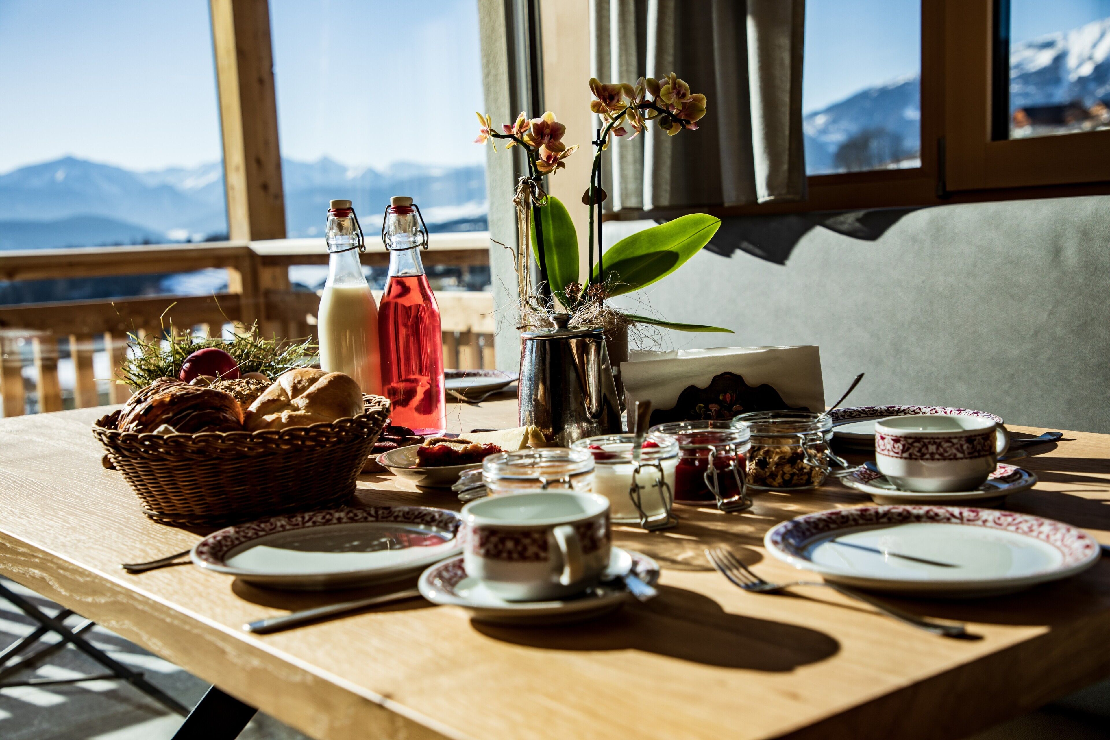 Una mesa de comedor con platos, tazas y una cesta de pan, frente a una ventana con vistas a la montaña.