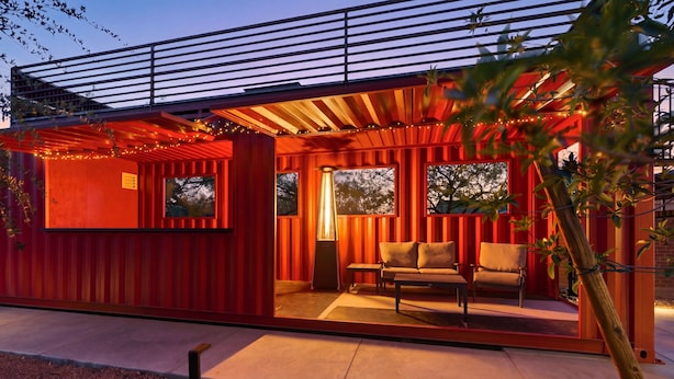Ein modernes Gebäude mit roter Fassade, einem überdachten Außenbereich mit Sofa und Tisch sowie großen Fenstern.