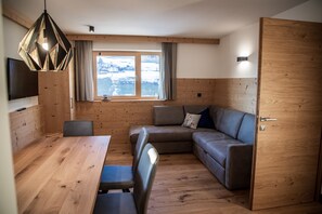 Une pièce aux murs lambrés, avec une table en bois, deux chaises, un canapé, un téléviseur et une fenêtre donnant sur des montagnes enneigées.