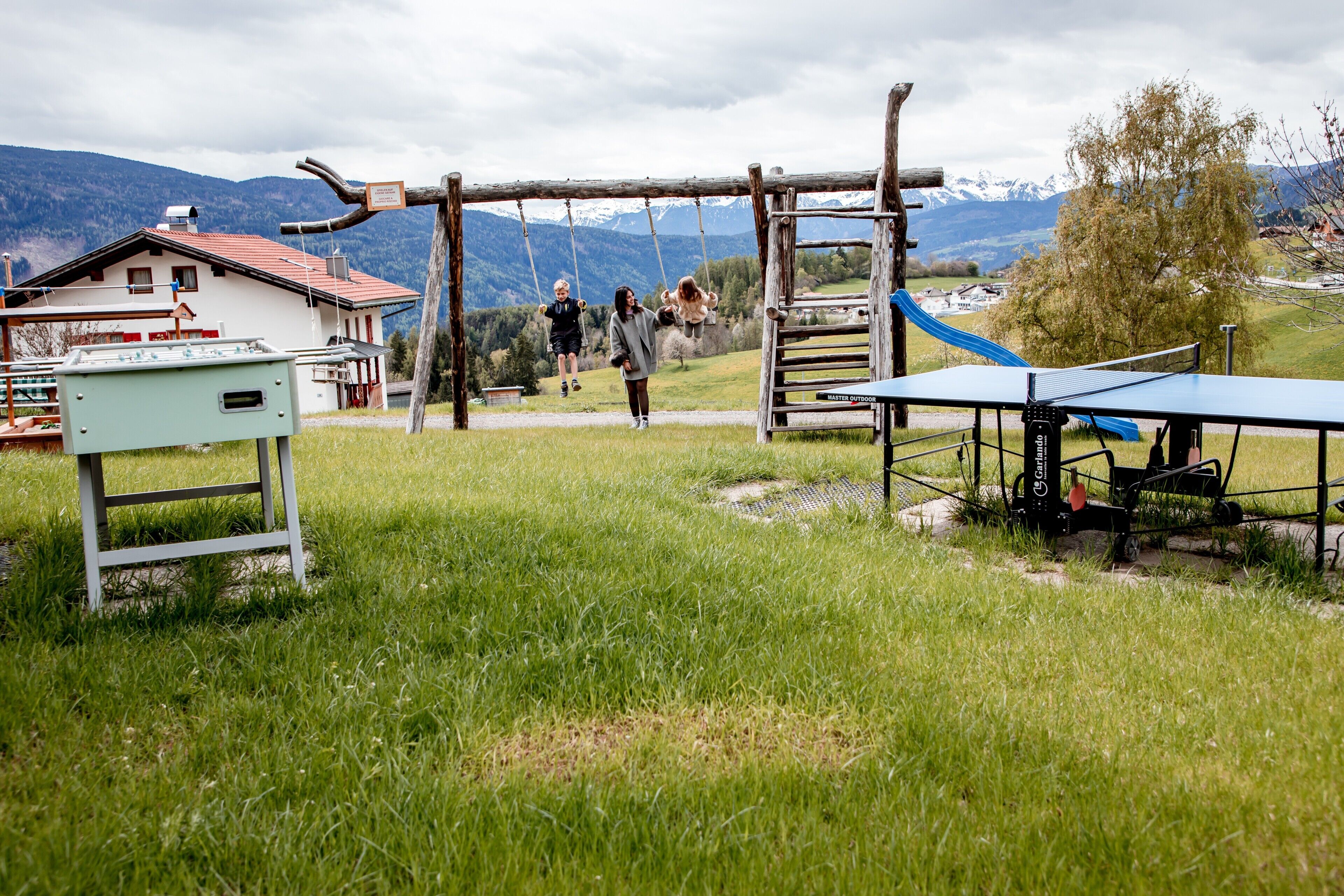 A playground with swings and a slide, a building with a red roof, and a mountain backdrop.