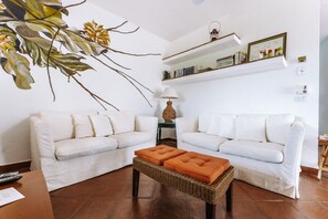 Un salon avec deux canapés blancs, une table basse en osier, une lampe sur pied et une étagère murale ornée de livres et d’objets décoratifs.