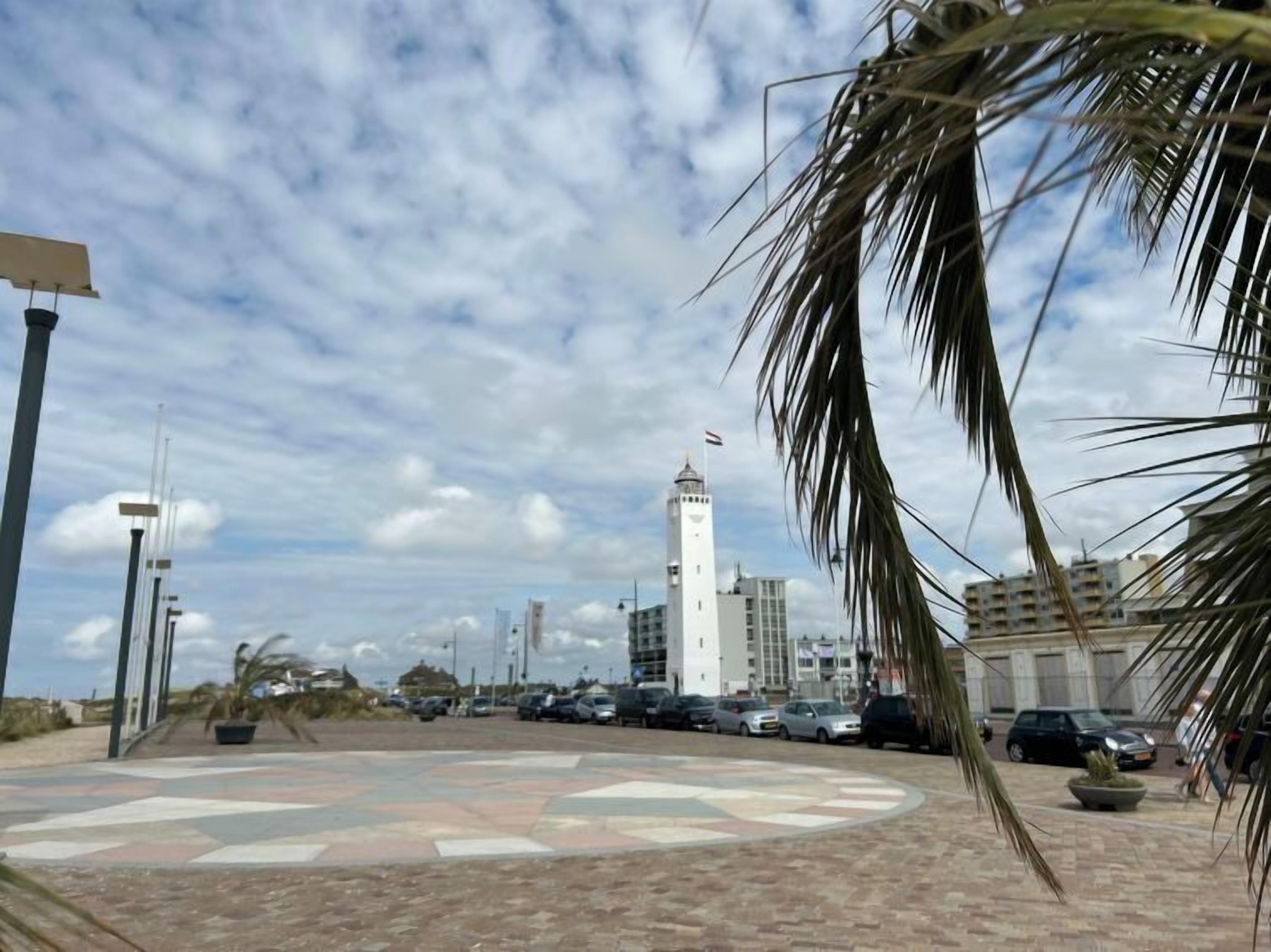 Una plaza con pavimento de diseño circular, una torre blanca alta con una bandera y coches estacionados.