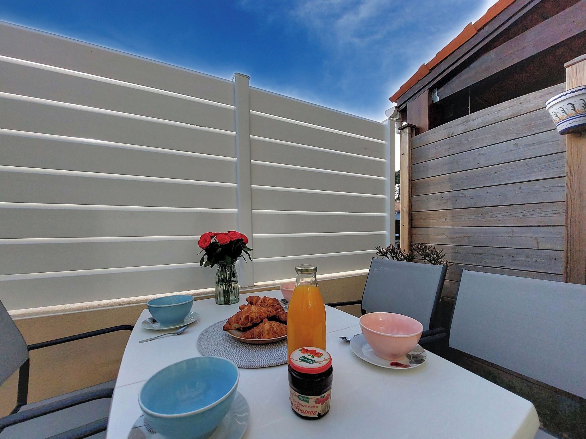 A table set outdoors with a white tablecloth, blue bowls, a pink bowl, a bottle of orange juice, a jar of jam, and a vase with red flowers.