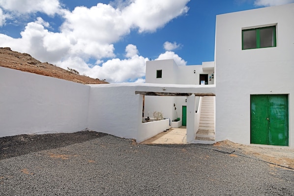 Un bâtiment moderne aux murs blancs, avec une porte et une fenêtre vertes, une cour recouverte de gravier et un ciel bleu dégagé.