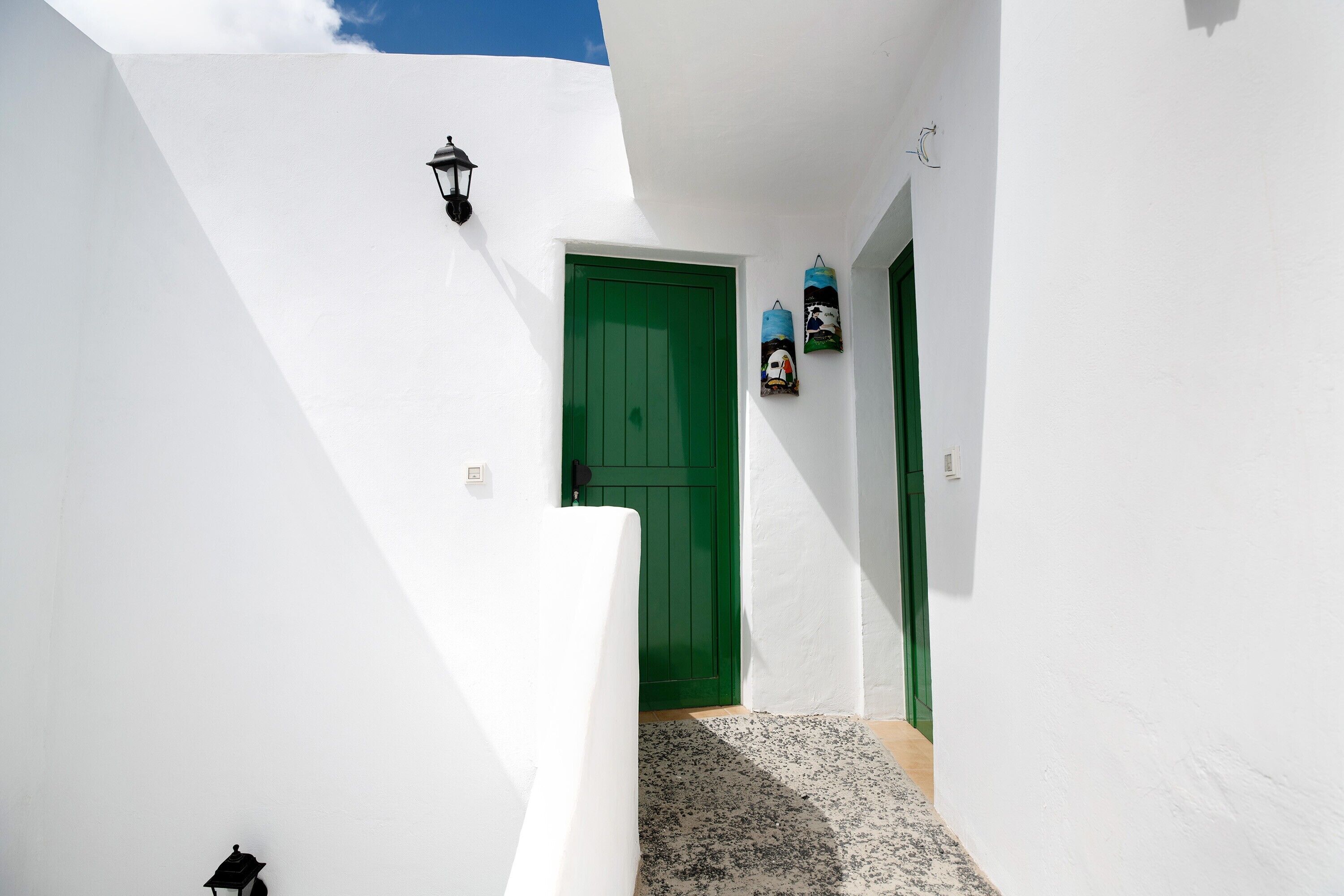 Un edificio de paredes blancas con una puerta verde, un pequeño escalón que lleva a la entrada y una lámpara fijada a la pared.