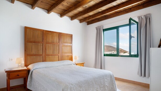 A bedroom with a wooden headboard, a bed with a white bedspread, a nightstand with a lamp, and a window with curtains.
