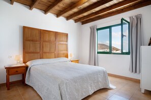 Une chambre à coucher avec une tête de lit en bois, un lit recouvert d’une courtepointe blanche, une table de chevet avec une lampe et une fenêtre avec des rideaux.
