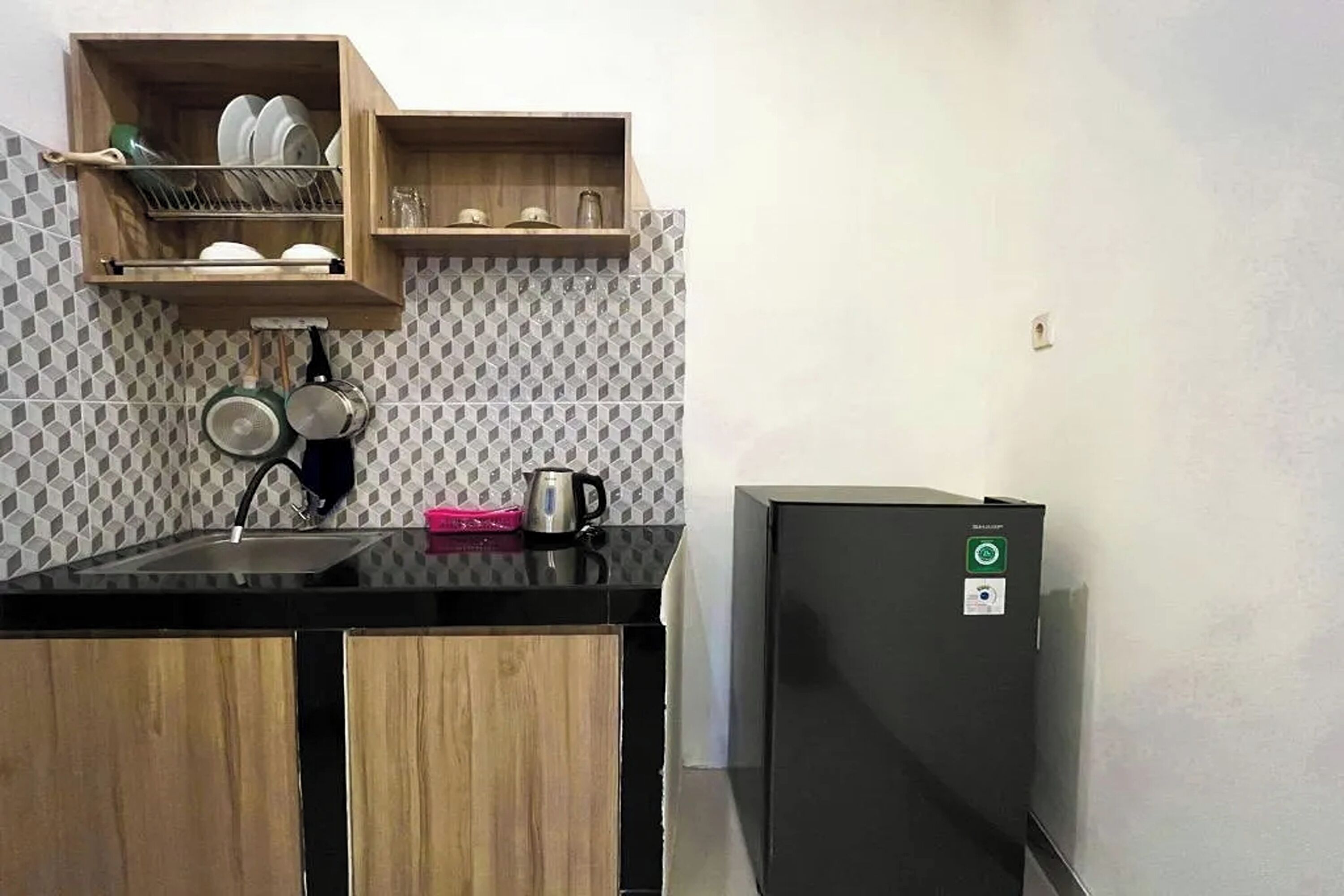 Eine kleine Küche mit schwarzer Arbeitsplatte, Holzschränken, einer eingebauten Ablage mit Geschirr, einem Wasserkocher und einem Kühlschrank.