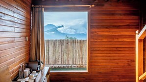 A wooden cabin with a wooden table set with cups and bottles, a window with a view of mountains and clouds, and a wooden fence outside. - The Dewi Eco Bamboo Villa (Kintamani)