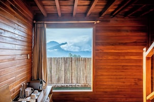 A wooden cabin with a wooden table set with cups and bottles, a window with a view of mountains and clouds, and a wooden fence outside.