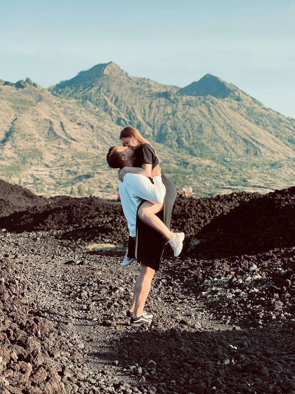 A couple embracing on a rocky terrain with a mountain in the background.