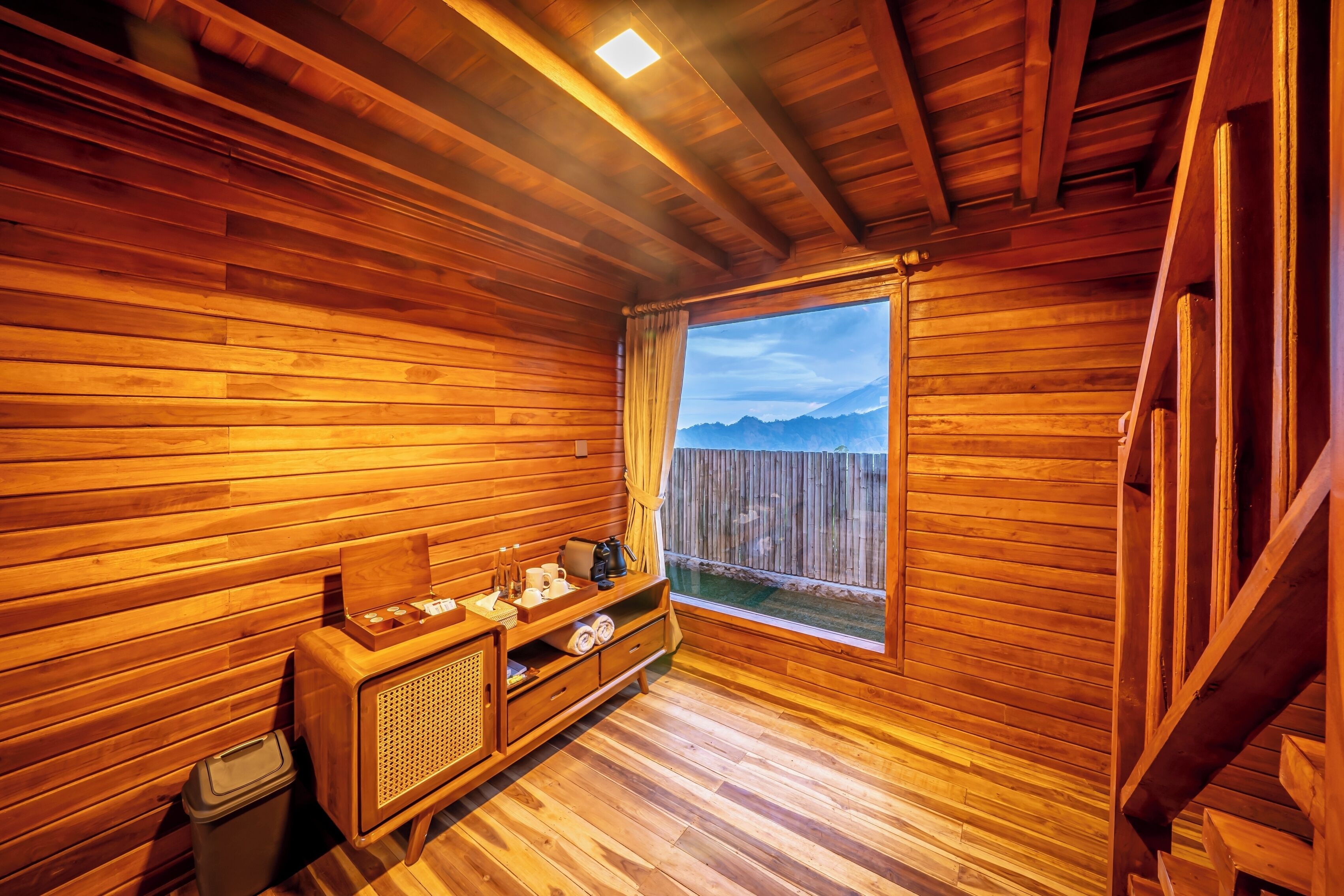 A wooden cabin with a wooden floor, a window with a view of mountains, a wooden cabinet, and a trash bin.