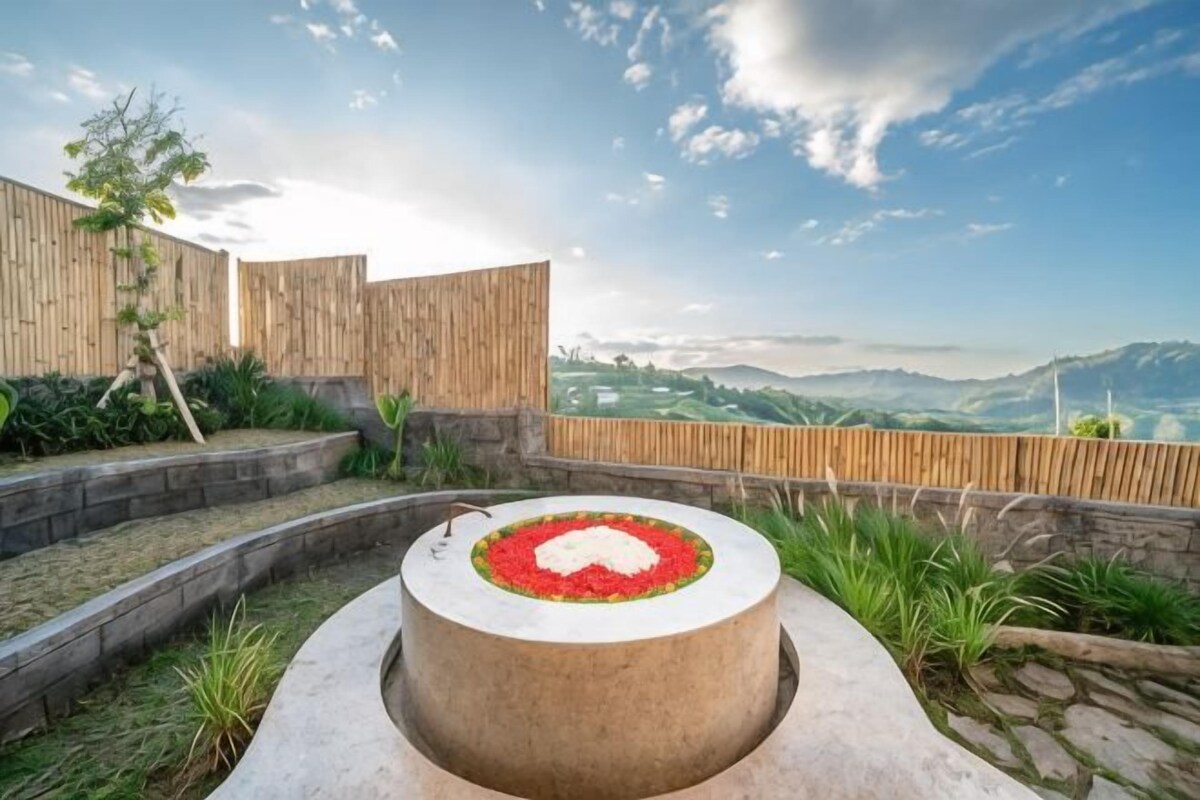 A circular stone structure with a flower arrangement, surrounded by a wooden fence and greenery, overlooking a mountainous landscape.