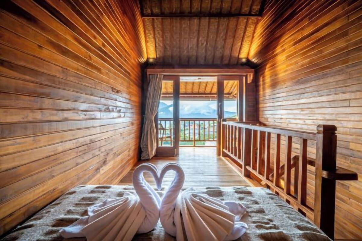 A wooden cabin interior with a cozy bed, a wooden railing, and a view of mountains through a glass door.