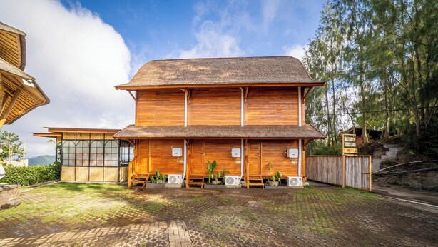 A two-story wooden building with a thatched roof, surrounded by a paved area with a brick pattern and greenery. - The Dewi Eco Bamboo Villa (Kintamani)