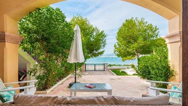 A patio with wicker furniture, a table with a book, and a view of a pool and trees. - Apartment 'Casa Faro' with Sea View, Wi-Fi and Air Conditioning (Puerto Pollensa)