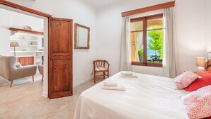 A bedroom with a bed, a chair, a wooden wardrobe, a window with curtains, and a view of greenery outside. - Apartment 'Casa Faro' with Sea View, Wi-Fi and Air Conditioning (Puerto Pollensa)