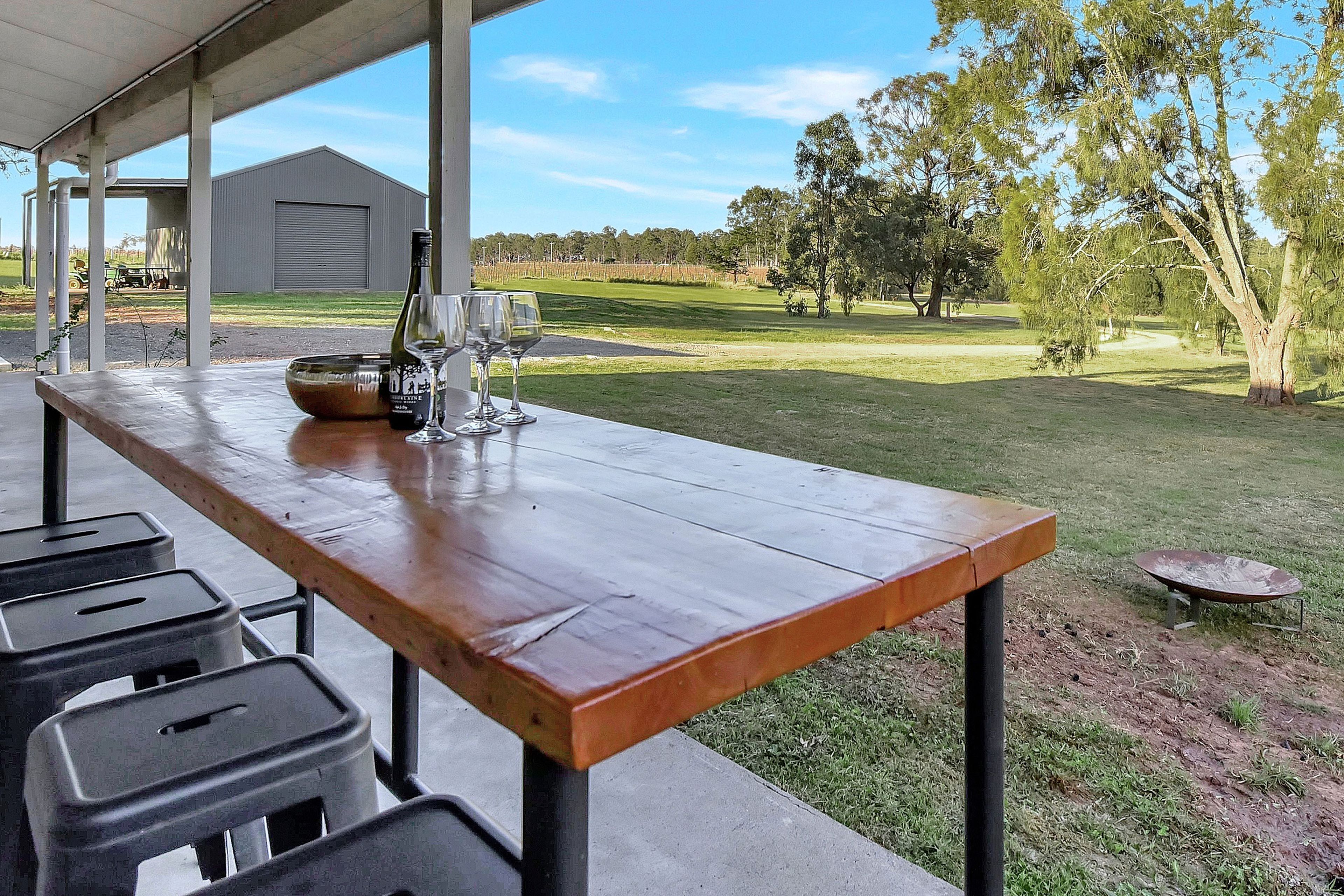 An outdoor dining area with a long wooden table, black chairs, and a bottle of wine.
