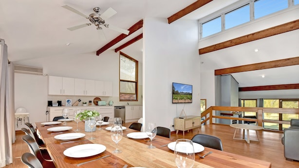 A spacious dining area with a long wooden table set for a meal, a kitchen in the background, and a large window providing natural light. - The Reserve Homestead walk to vineyards (Pokolbin)