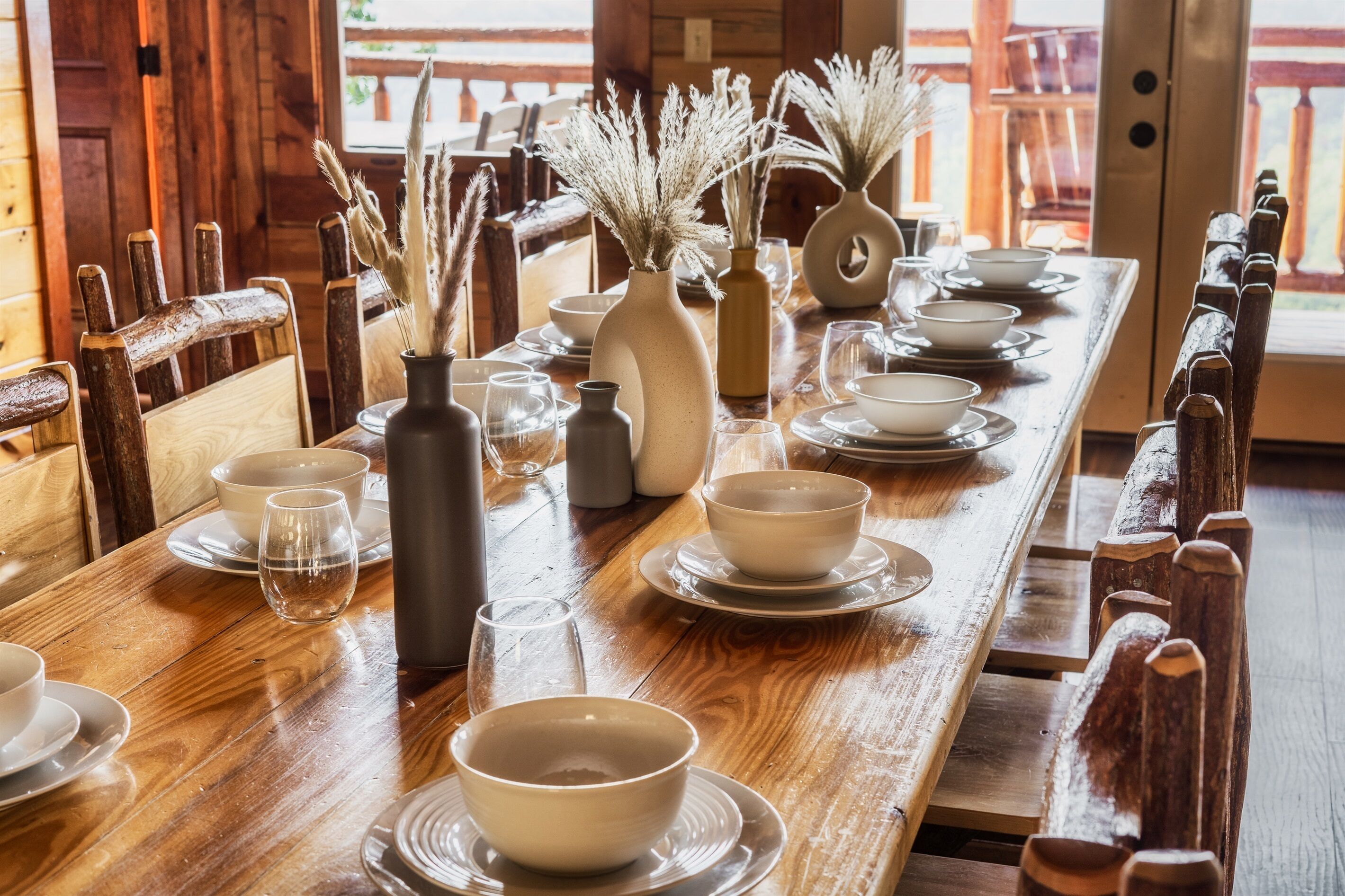 A wooden dining table set with white bowls, plates, and glasses, accompanied by decorative vases with dried plants.