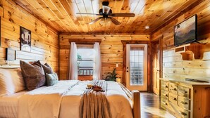 L’intérieur d’une cabane en bois, avec un lit, un ventilateur de plafond, un téléviseur et une fenêtre avec des rideaux.