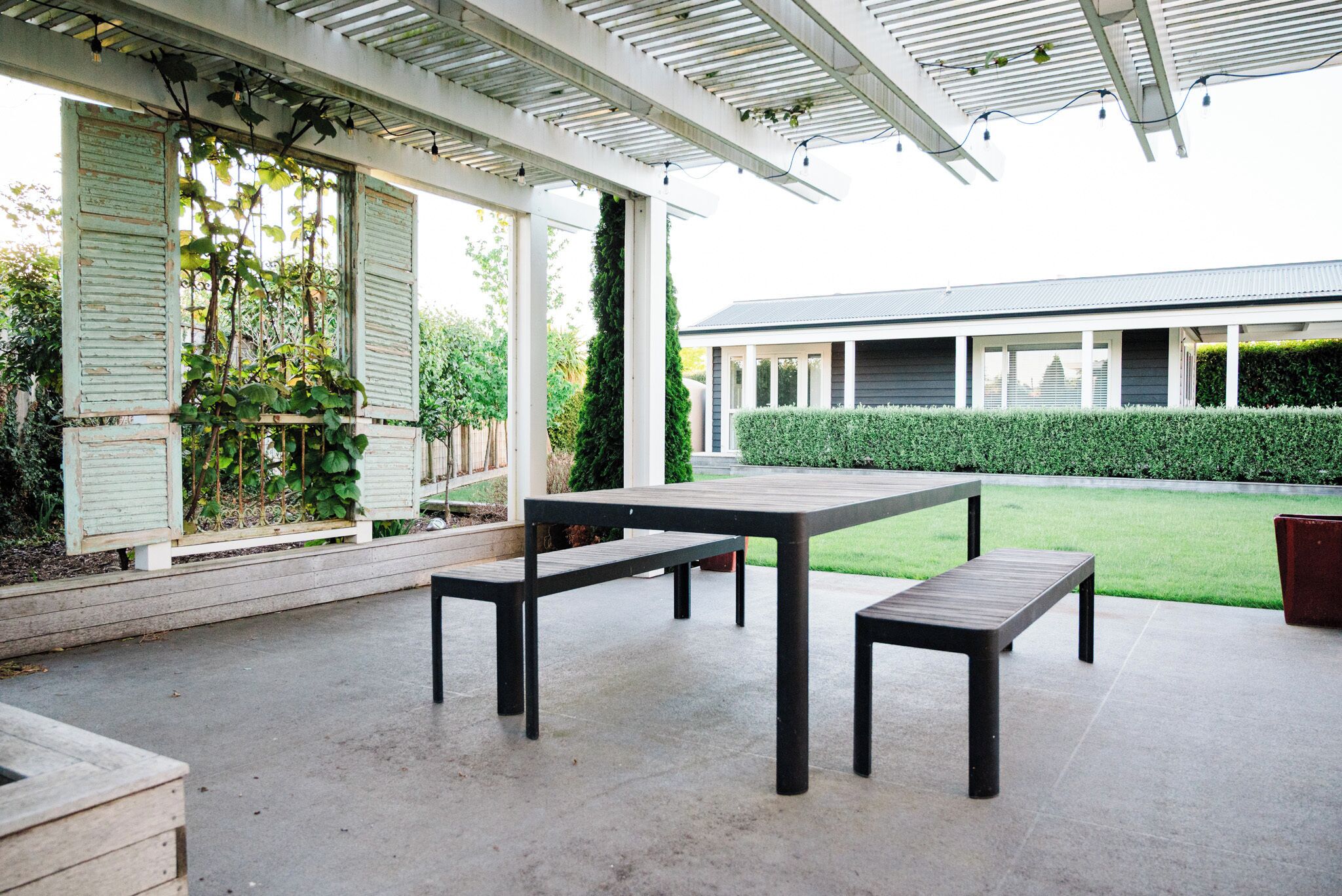 An outdoor patio with a table and benches, a covered area with a pergola, and a building in the background.