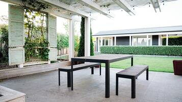 An outdoor patio with a table and benches, a covered area with a pergola, and a building in the background.