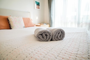 A neatly made bed with rolled towels, a pillow, and a bedside table with a book and a lamp.