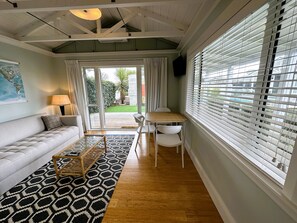 A modern living room with a sofa, a coffee table, and a dining table with chairs. There is a large window with blinds, a lamp, and a view of the outdoors.