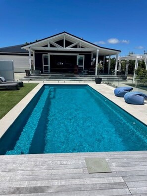 Uma piscina retangular com um deck de madeira na frente, uma área de varanda coberta com assentos e uma casa ao fundo.