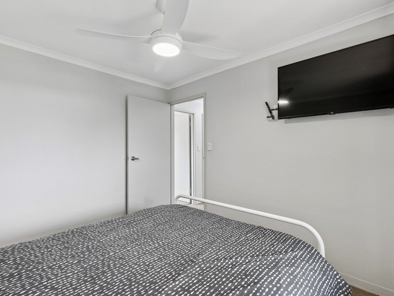 A bedroom with a bed, a ceiling fan, a TV mounted on the wall, and a door leading to another room.