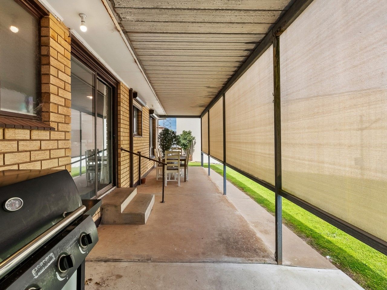 A covered outdoor area with a barbecue grill, a concrete pathway, and a view of a building with a brick facade.