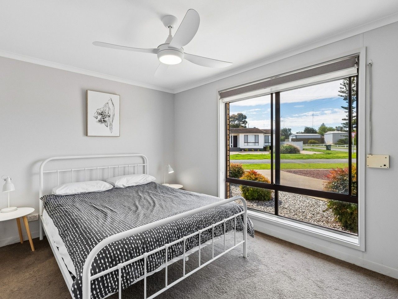 A bedroom with a bed, a ceiling fan, a window with a view of the outside, and a small bedside table.