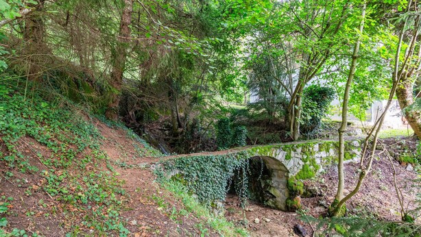 Une zone boisée avec un pont de pierre, un sentier de terre et un mur de pierres.