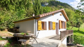 Un bâtiment blanc avec des volets jaunes, une terrasse avec une table et des chaises, et un escalier en pierre menant à une plateforme en bois.