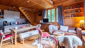 L’intérieur d’un chalet en bois comprend une cuisine, un coin repas et un salon, ainsi qu’une chambre en mezzanine et de grandes fenêtres.