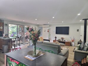 Una sala de estar moderna con comedor, televisor montado en la pared y un ventanal con vistas a un jardín.