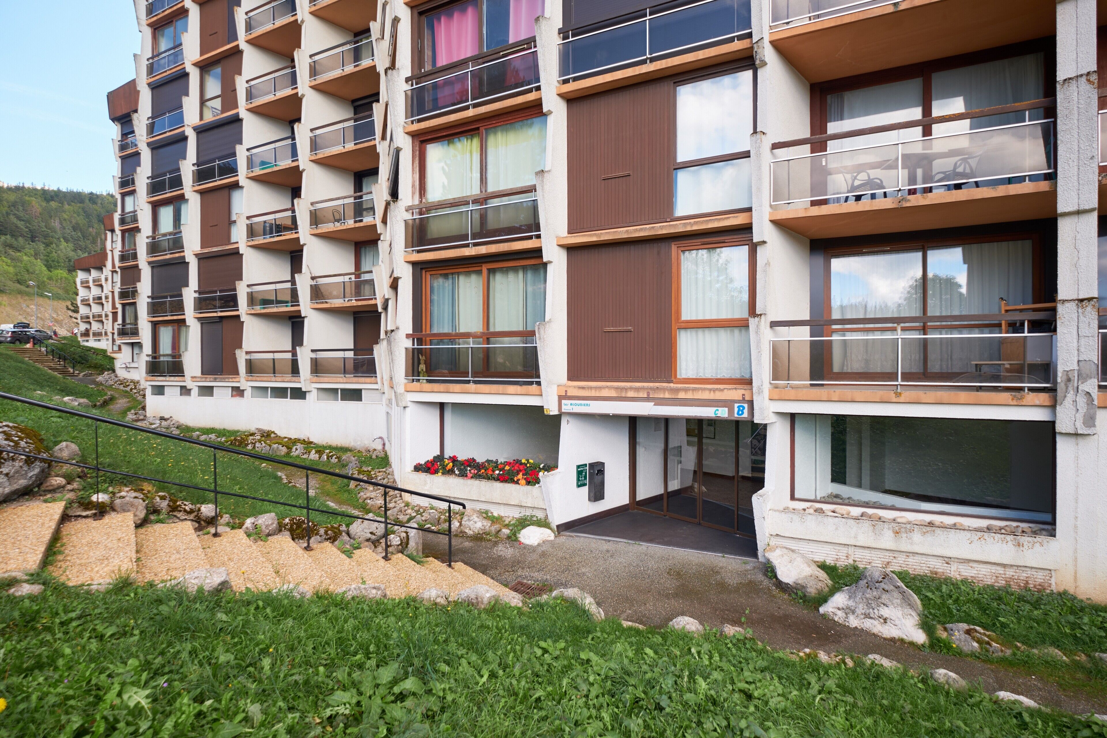 A multi-story hotel with balconies, a staircase, and a flower bed.