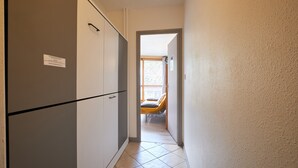 En smal hallgång med en säng längre bort, en dörr på vänster sida och ett klinkergolv.