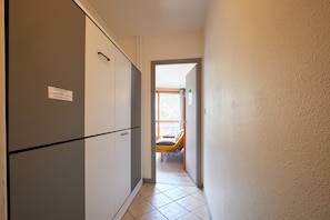 Un couloir étroit, avec un lit visible au fond, une porte sur la gauche et un sol carrelé.