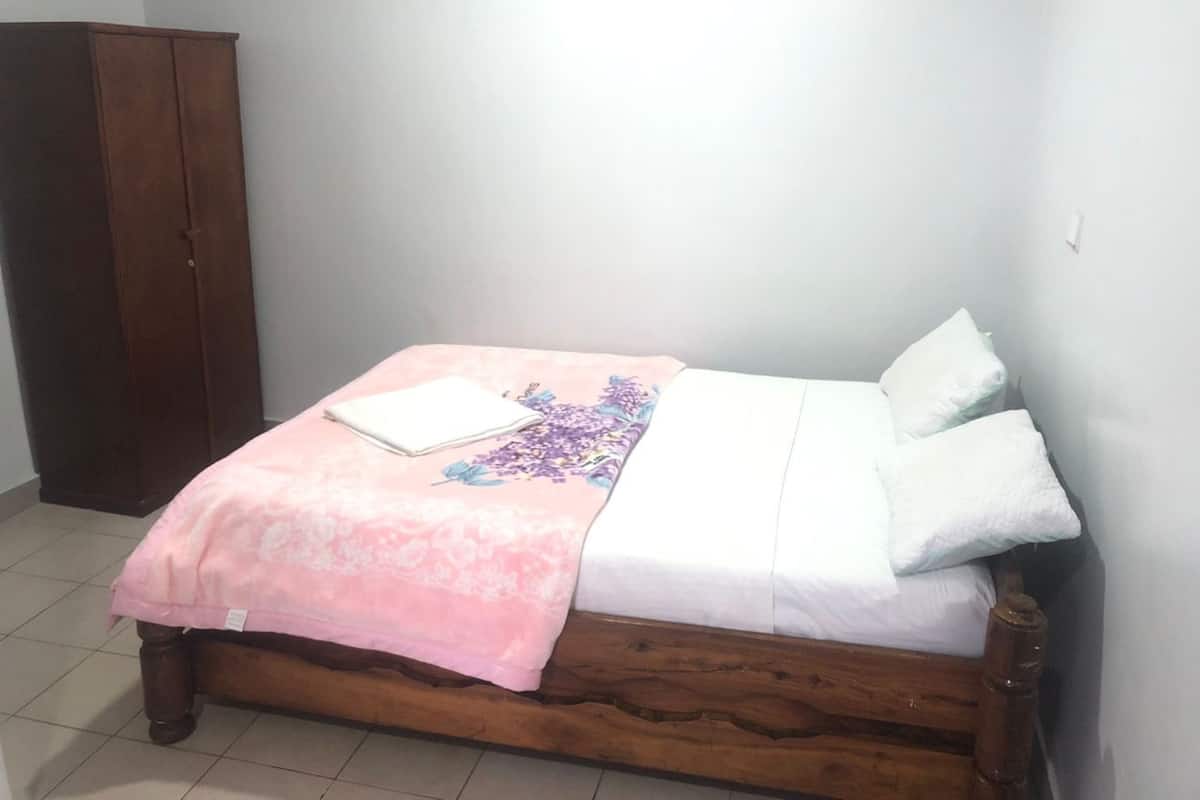 Un lit simple avec une couvre-lit à motifs floraux, un oreiller blanc et un vêtement blanc plié posé sur le lit. Une armoire en bois se trouve à gauche du lit.