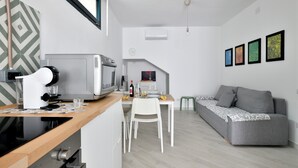 A modern kitchen with a microwave, stove, and coffee machine, a dining area with a white table and chair, and a grey sofa in a living area with a staircase. - Vacation Home "Appartamentino" with Private Terrace & Wi-Fi (Barisardo)