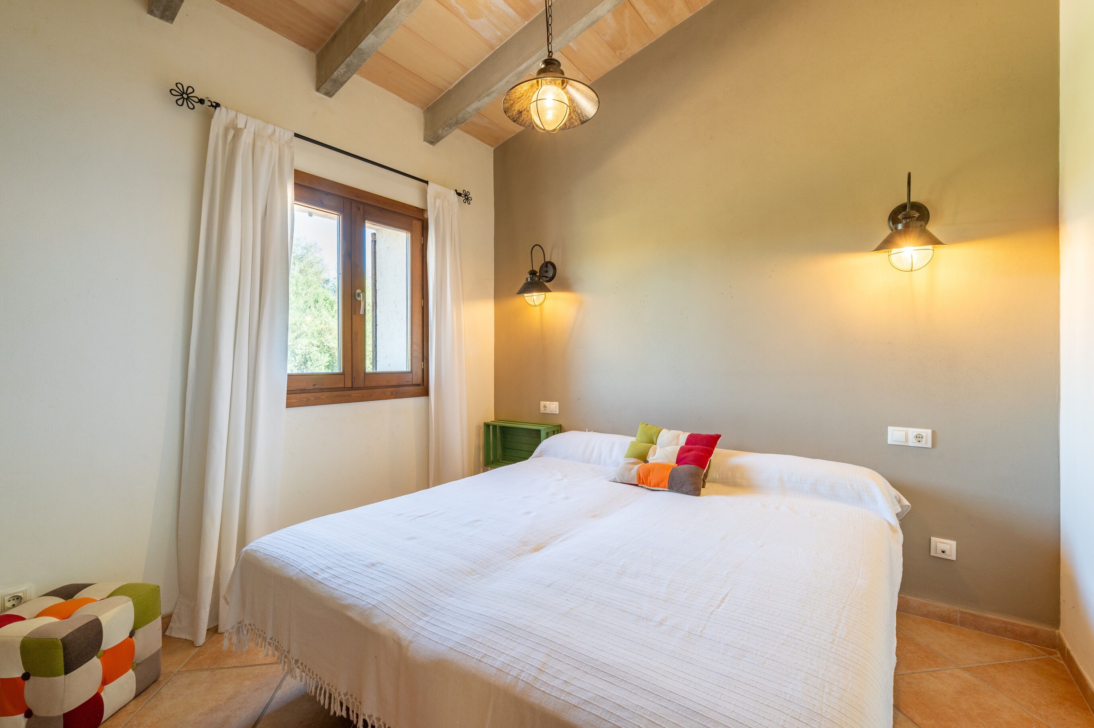 A bedroom with a bed, a window with curtains, a hanging light fixture, and a wall-mounted light.