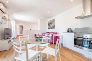 Una cucina moderna con una cappa in acciaio inossidabile, una zona pranzo con tavolo in vetro e sedie bianche, e un divano rosa nella zona giorno.